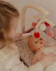 Kislány babaágyban pelenkázó babával, felette pomponos fa babajáték