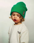 Gyermek zöld kötött sapkában, fehér pulóverben, világos háttérrel.