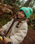 Mosolygó kisgyerek zöld kötött sapkában, puha bézs kabátban, erdei környezetben játszik.