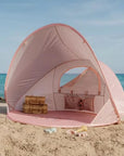 Rózsaszín strandsátor homokos tengerparton, gyerek játékokkal, kék tengerrel a háttérben.