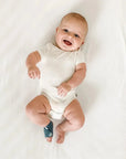 Owlet Dream Sock - Mosolygó kisbaba fehér rugdalózóban, okos zoknival a bal lábán, fehér ágyon fekve