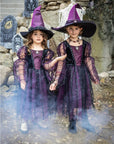 Két kislány boszorkány jelmezben, lila fekete ruhában, halloween dekorációval a háttérben