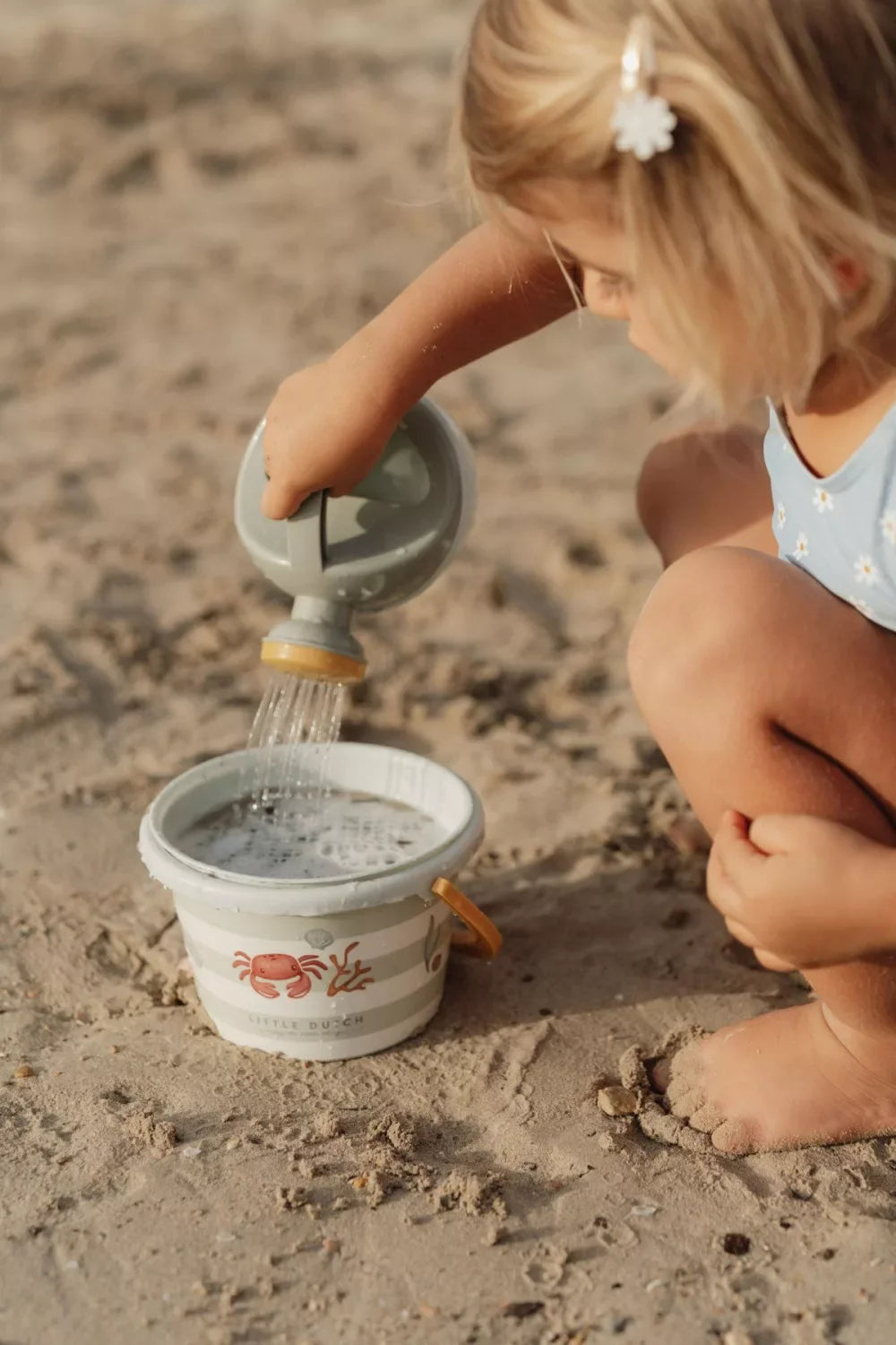 Kisgyermek játszik a homokban, homokozó vödörbe vizet önt, strand játék