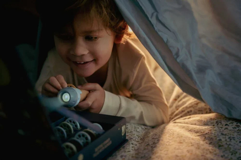 Mosolygó kisgyerek játszik zseblámpával ágyban, plédből sátor alatt