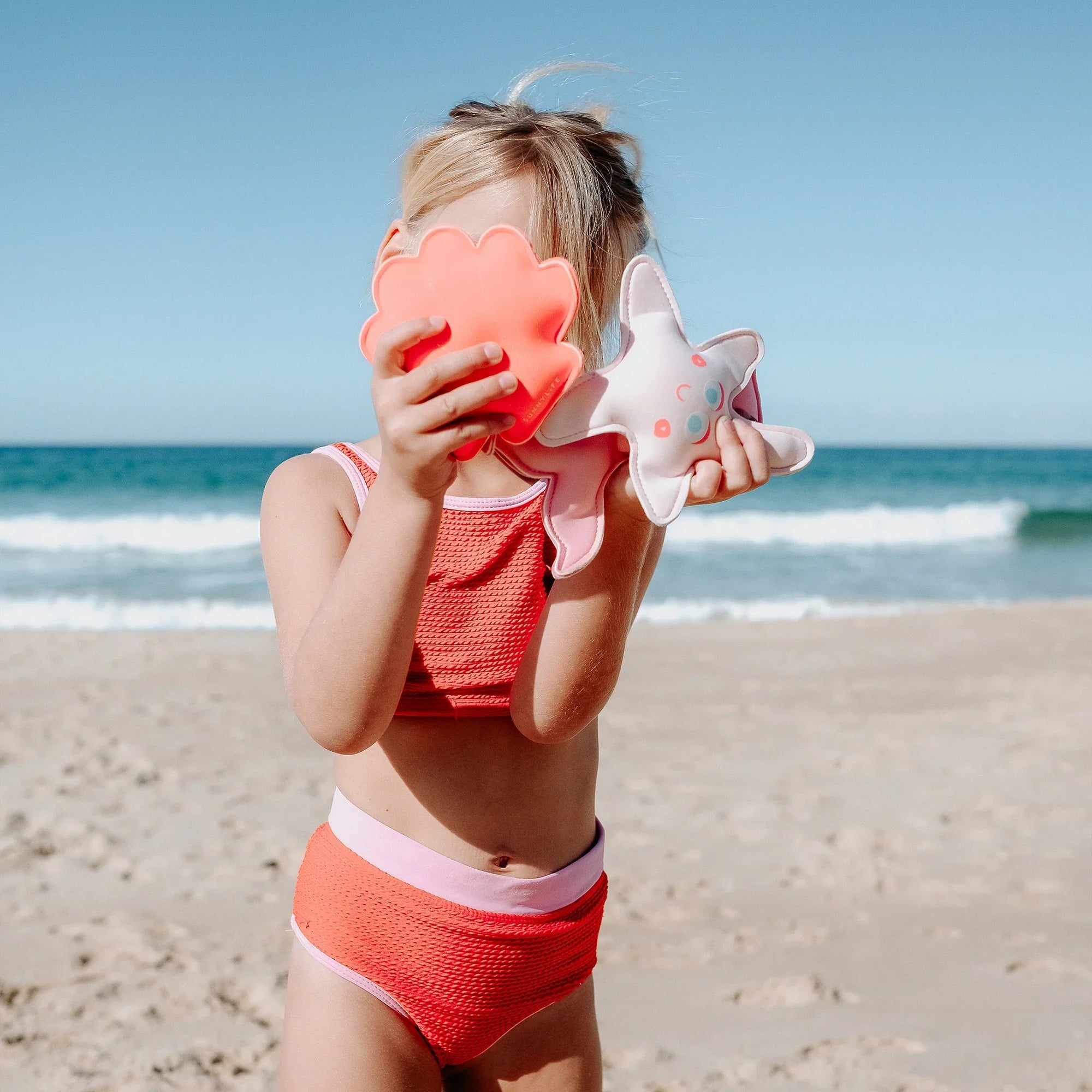 Kislány rózsaszín fürdőruhában, tengeri csillag és kagyló formájú strandjátékkal a tengerparton