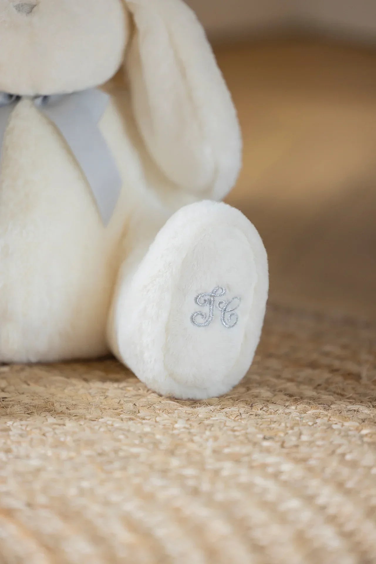 Fehér plüss nyuszi szürke masnival és hímzett tappancson, fonott szőnyegen.